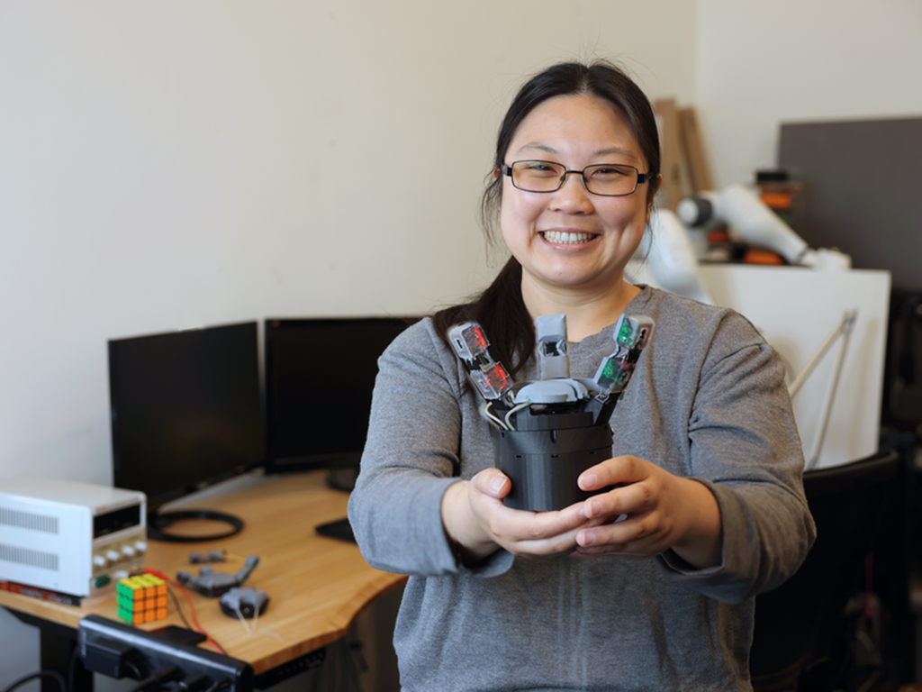 Sandra Liu poses for the camera holding her GelPalm prototype, a robotic hand with sensors. She is in a lab workspace with two computer monitors, a Rubik's cube, and electronic equipment.