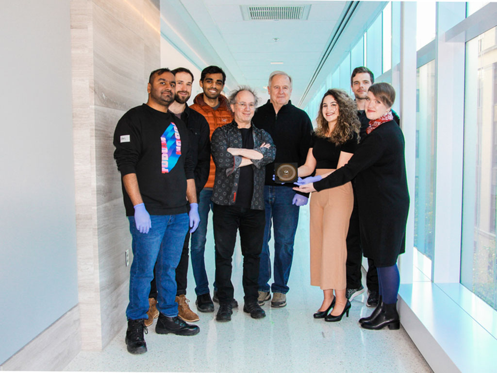 Eight people pose standing in a white hallway with the HUMANS nanowafer held between them. On their left is a wall, and the right are windows.