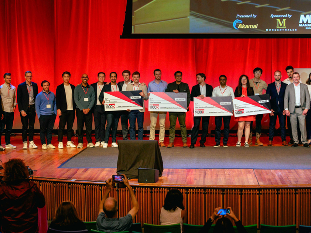 21 young professionals on stage with a few holding ceremonial check prizes, the MIT 100k Competition awards