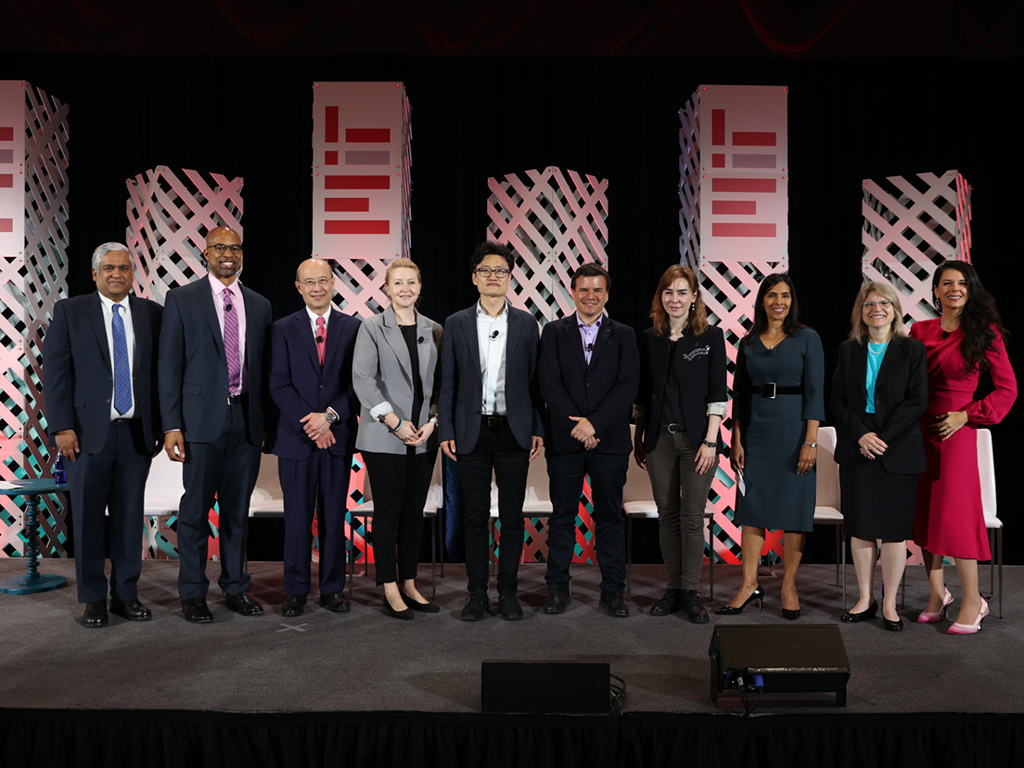 Participants in the panel discussion stand side-by-side on the stage, posing for group photo. Square columns with the MIT logo rise up behind them.