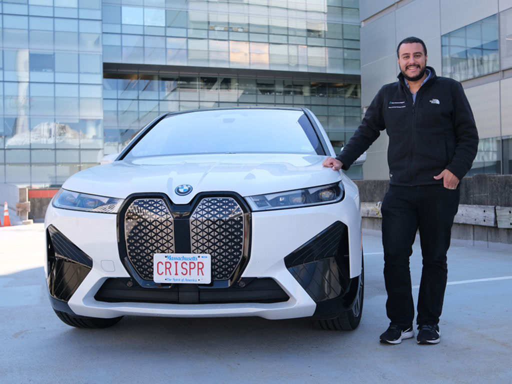 Omar Abudayyeh stands on a rooftop next to a white BMW SUV with the license plate, “CRISPR.” MIT lab buildings are in the background.