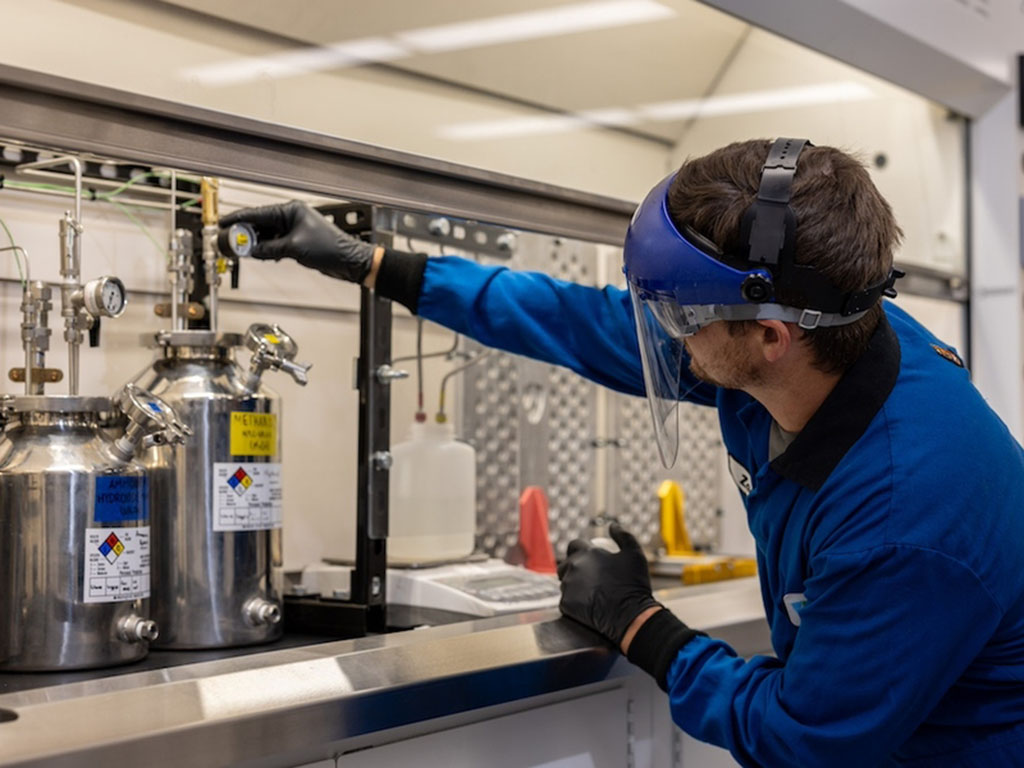 Zach Goodman, wearing full lab protection, adjusts a gauge above a stainless steel cylinder in a lab